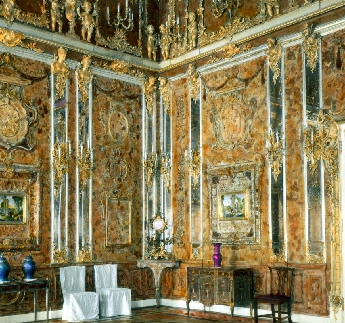 A corner of a palace room faced floor to ceiling in mottled amber panels framed by gilded carving, with tall mirrored pilasters, gilded cherub figures along the cornice, two framed landscape mosaics, a marquetry commode, and two chairs under white dust covers.